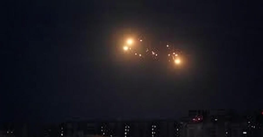 A vibrant display of fireworks exploding in the night sky with bursts of red, orange, and white, viewed from a distance with silhouettes of people watching and a cityscape with illuminated buildings below.