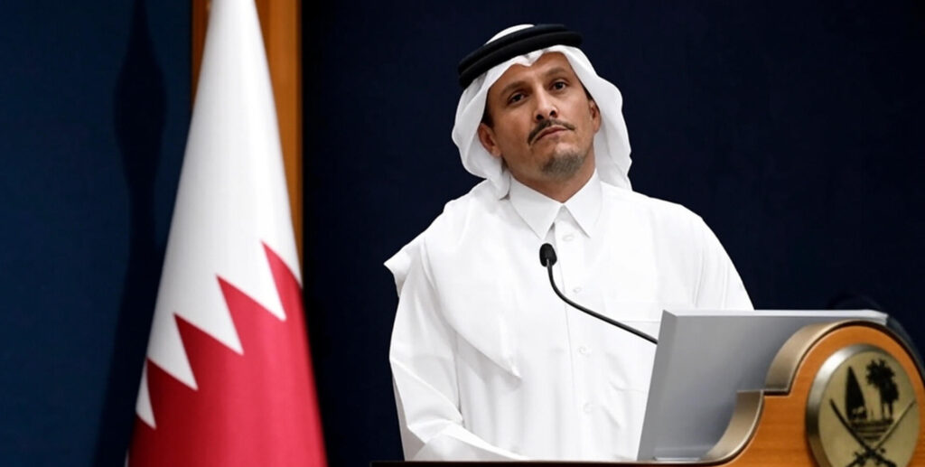 The image shows a man wearing traditional white robes and a headscarf, standing at a podium with a microphone. The podium features an emblem with crossed swords and a palm tree. To the left of the man, a flag with a white background and a red vertical stripe with a serrated edge is visible against a dark blue backdrop.