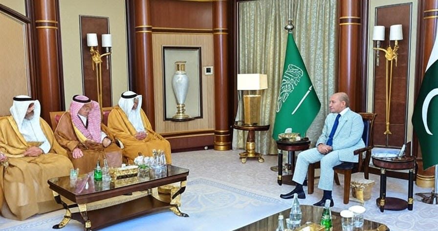 Two men walking side by side in a spacious, well-lit hallway with marble floors and large windows. The man on the left is wearing a gray double-breasted suit with a blue shirt and a polka-dot tie, while the man on the right is dressed in a traditional white robe with a gold trim and a headscarf. Another man in a similar white robe follows behind them.