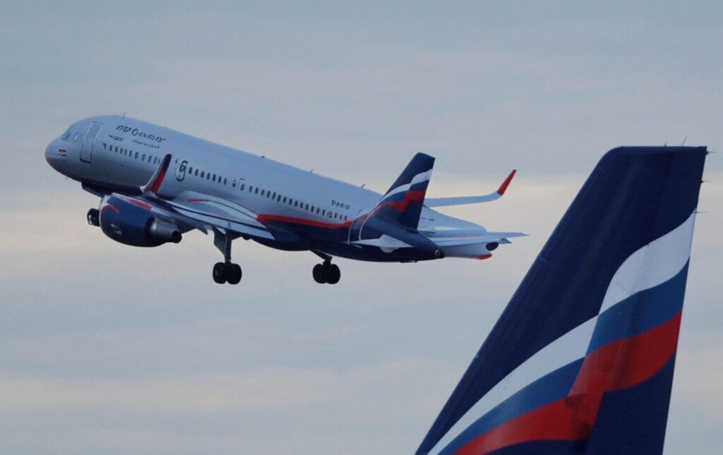 The image depicts an airplane in flight, captured from a low angle with its landing gear extended, suggesting it is either taking off or landing. The aircraft features a livery with the word "Аэрофлот" (Aeroflot) written on the fuselage, indicating it is operated by Aeroflot, the Russian national airline. The tail of the plane displays a design incorporating the colors of the Russian flag: blue, red, and white. In the foreground, part of another aircraft's tail fin with a similar color scheme is visible, reinforcing the Aeroflot branding. The background consists of a cloudy sky, providing a neutral setting for the aircraft.