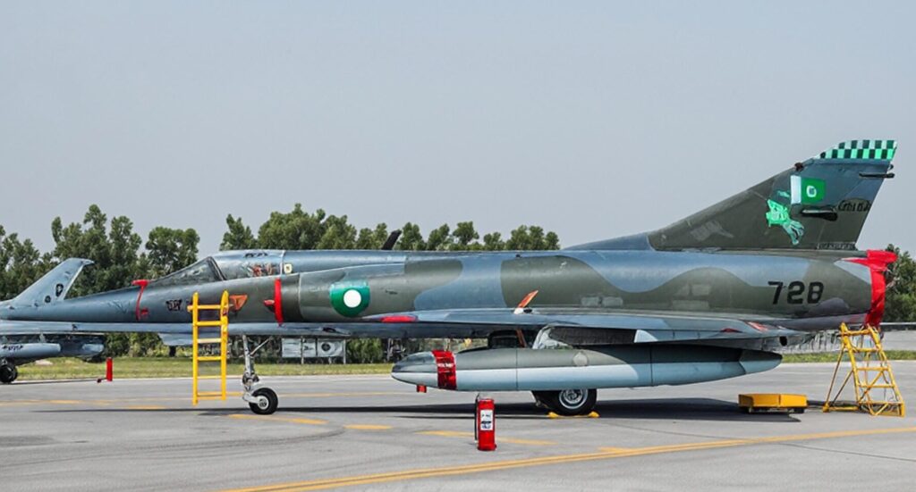Military aircraft with Pakistani markings, including a green and white flag and a rearing horse emblem, parked on a tarmac with yellow ladders and a red fire extinguisher nearby, trees in the background.