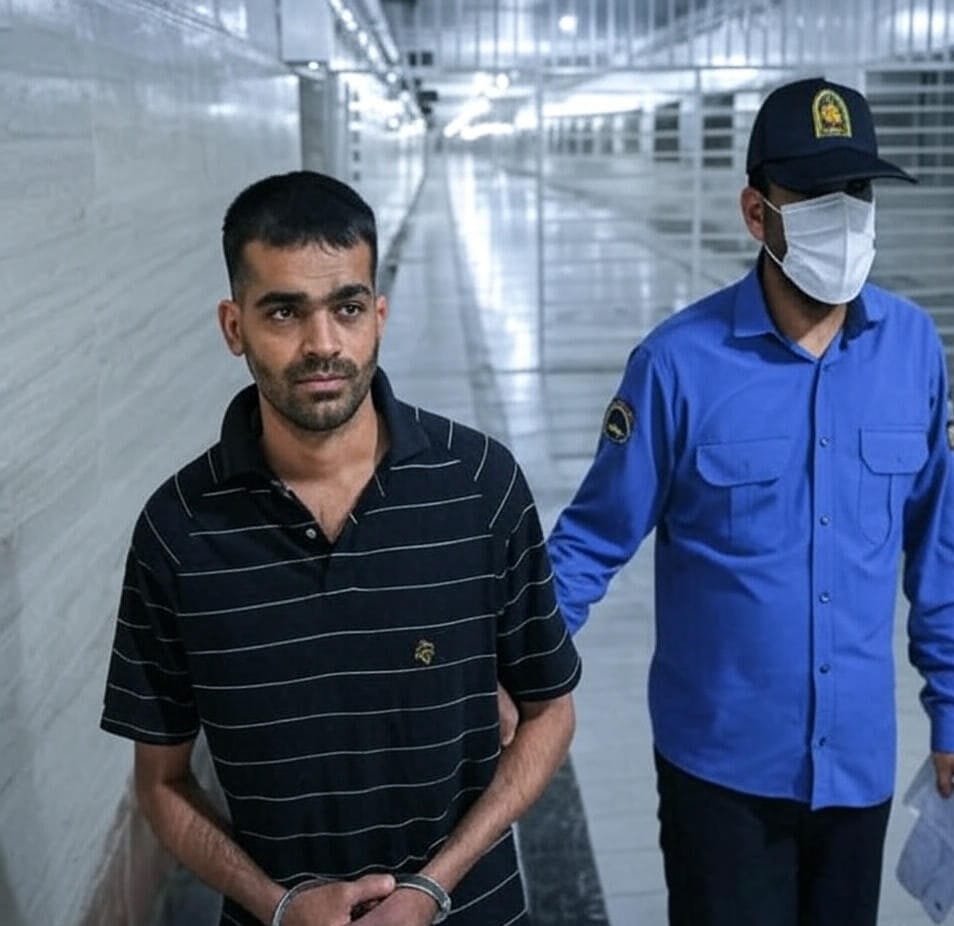 The image depicts a scene inside a hallway with a tiled wall and a series of doors or cells on the left side. A man in a dark, short-sleeved, striped polo shirt with his hands handcuffed in front of him stands in the foreground. He appears to be a prisoner or detainee. To his right, a uniformed officer wearing a blue shirt, dark pants, a cap with an emblem, and a face mask is holding the man's arm, escorting him. The hallway is well-lit with a modern, institutional appearance, suggesting a detention or prison facility.