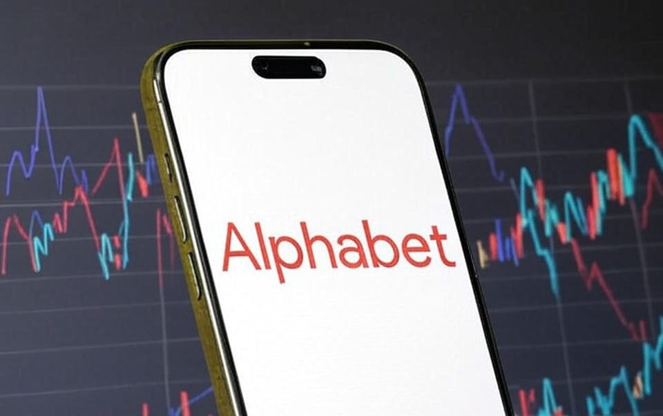 The image shows a smartphone displaying the word "Alphabet" in red text on its screen, held against a blurred background featuring a stock market chart with fluctuating lines in red, blue, and green. The setting suggests a financial or business context, with the focus on the Alphabet brand, the parent company of Google.