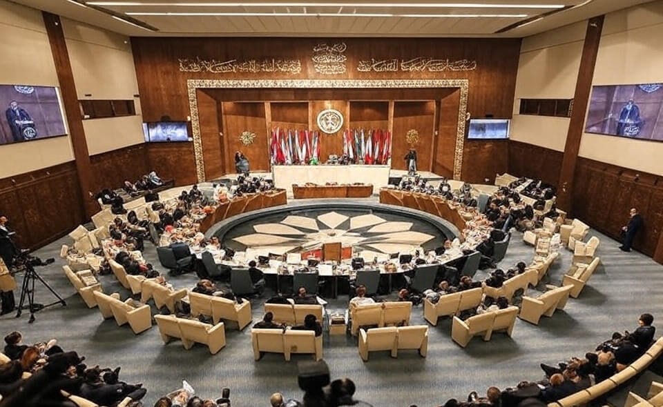 The image depicts a formal conference or assembly taking place in a circular chamber with a central table surrounded by delegates seated in a semi-circular arrangement. The room features ornate wooden paneling, a large emblem on the wall, and multiple national flags displayed at the front. Two large screens on either side of the room show a speaker addressing the assembly. The audience, seated in rows of cushioned chairs, includes individuals taking notes and operating cameras, indicating a significant official event.