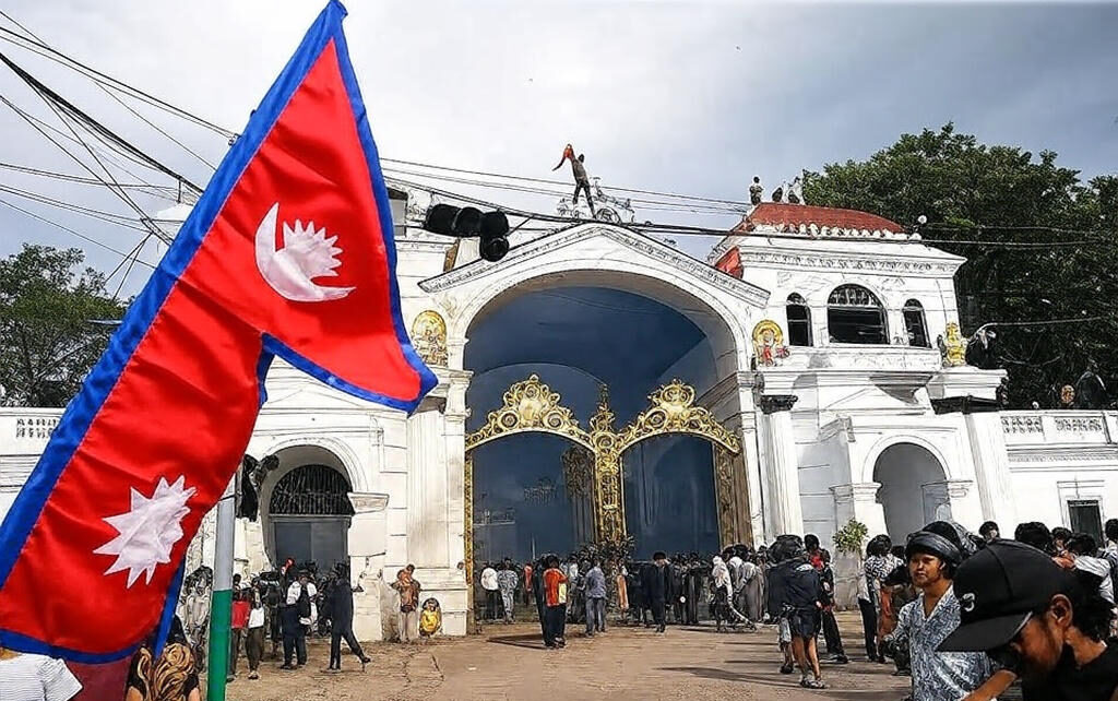 The image depicts a tense protest scene in front of a grand, ornate gate with golden decorations, part of a significant building in Kathmandu, Nepal. A large Nepalese flag, featuring red, blue, and white colors with a sun and crescent moon emblem, is prominently displayed in the foreground, held by a protester. In the background, a person is seen climbing onto the gate's structure, waving a flag, while a crowd of demonstrators gathers, some holding additional flags and others observing. Smoke and flames rise from a fire on the ground, adding to the chaotic atmosphere, with power lines crisscrossing above and a cloudy sky overhead.