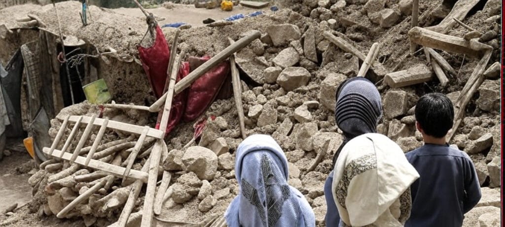 The image depicts a scene of destruction with a pile of rubble, including broken wooden beams and stones, likely from a collapsed structure. Three individuals, dressed in traditional clothing with headscarves, are seen observing the debris from a distance. In the background, there are remnants of a damaged building with a red fabric hanging loosely. The setting appears to be a rural area with earthen walls and scattered household items.