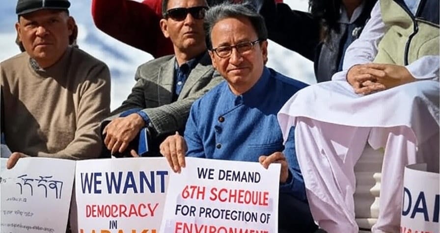 The image shows a group of individuals seated and holding signs during what appears to be a protest or demonstration. The central figure holds a sign that reads "WE DEMAND 6TH SCHEDULE FOR PROTECTION OF ENVIRONMENT" with "6TH SCHEDULE" and "FOR PROTECTION OF ENVIRONMENT" in bold. To the left, another person holds a sign stating "WE WANT DEMOCRACY IN LADAKH" with "DEMOCRACY" and "IN LADAKH" highlighted. Additional signs with text in a different script and the word "DAANDS" are also visible. The individuals are dressed in traditional and casual clothing, and the setting suggests an outdoor gathering.
