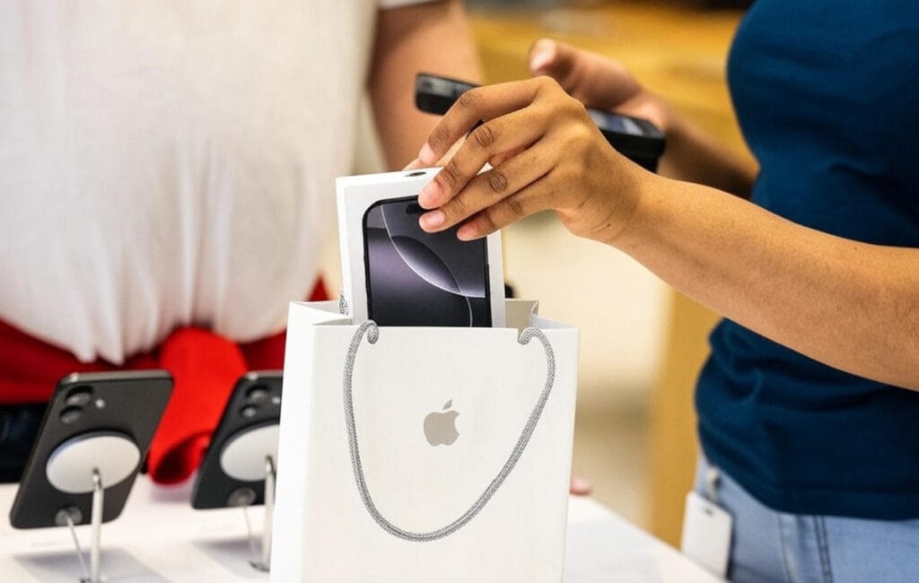 The image shows a close-up of a hand placing a smartphone box into a white Apple-branded shopping bag. The box contains a visible black smartphone with a circular camera design. The scene takes place on a counter displaying additional smartphones on stands, with a blurred background featuring a person wearing a red shirt and another individual in a blue top, possibly an employee, handling a payment device. The setting appears to be an Apple store.