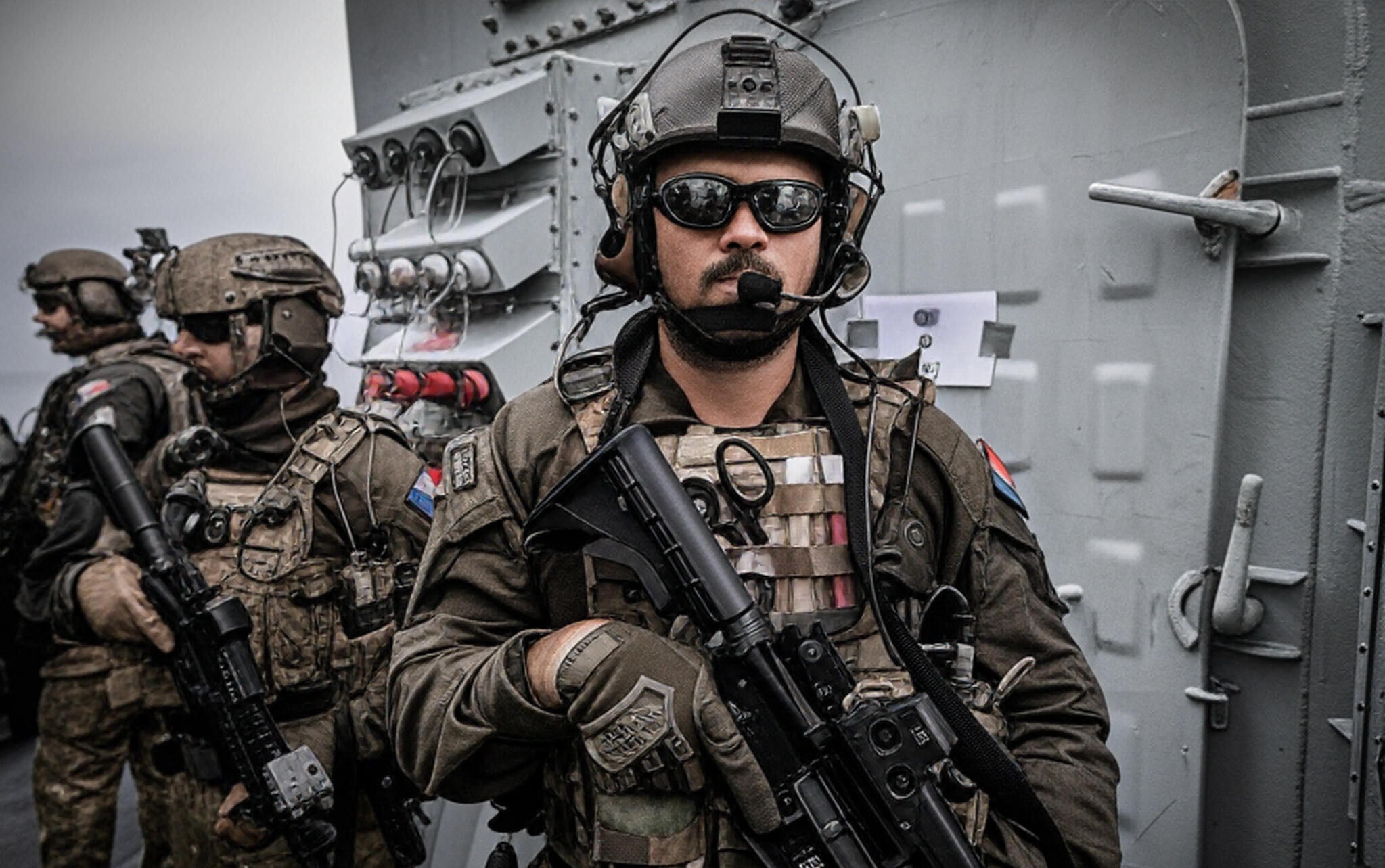 The image shows a group of military personnel in tactical gear, standing on what appears to be the deck of a naval vessel. The individual in the foreground is equipped with a helmet, night vision goggles, and a communication headset, holding a rifle with both hands. The uniform includes a tactical vest with various pouches and a patch featuring the colors of the Romanian flag. In the background, additional personnel are visible, similarly equipped, with a metallic hatch and control panel of the vessel behind them. A sign reading "OUT" is affixed to the hatch.