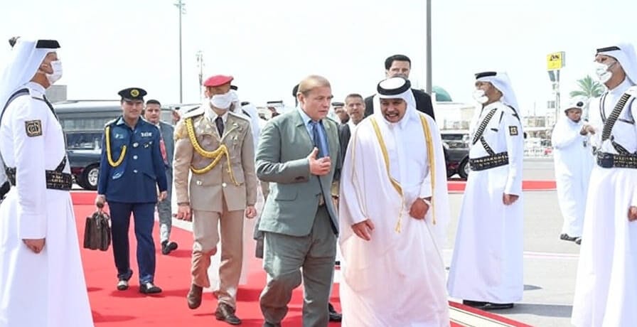 A group of dignitaries and uniformed officials walking on a red carpet, escorted by guards in traditional white robes and military attire, with vehicles and a modern building in the background.