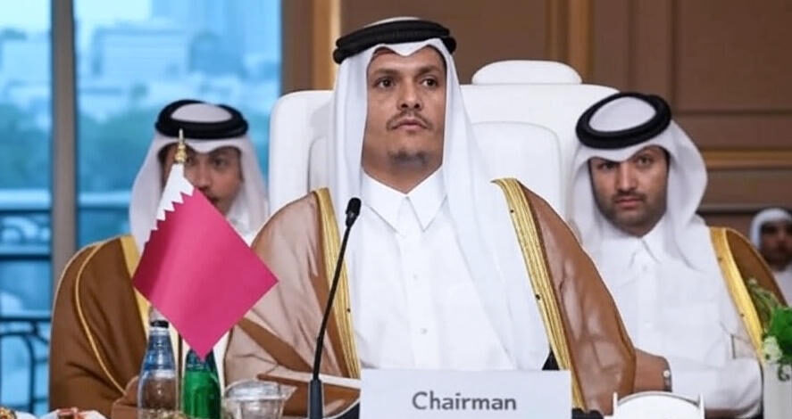 The image shows three individuals dressed in traditional Middle Eastern attire, seated at a formal meeting or conference table. The central figure is positioned behind a nameplate labeled "Chairman" and is flanked by two others. A microphone is placed in front of the central figure, and a small flag of Qatar is visible on the table. The table also holds water bottles, glasses, and other items, suggesting an official proceeding. The background includes a window with a view of a cityscape and a decorative interior setting.