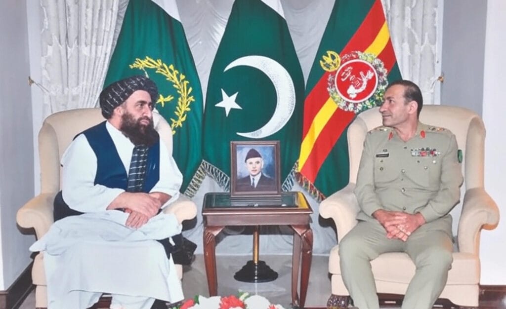 A diplomatic meeting between a Taliban representative in traditional attire and a Pakistani military official in uniform, seated with flags of Pakistan and Afghanistan in the background. A framed portrait and floral arrangement on a table symbolize ongoing efforts to address Afghanistan-Pakistan border tensions in 2025, as highlighted in regional mediation discussions.