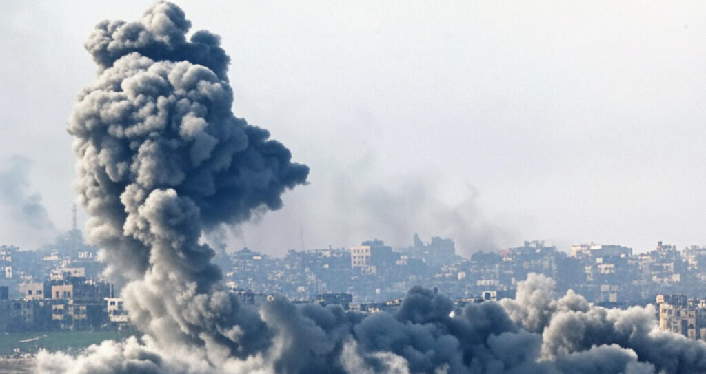 The image depicts a scene of significant smoke rising from multiple locations over a densely built urban area. Thick, dark clouds of smoke dominate the foreground, suggesting recent explosions or fires. The background features a hazy skyline of buildings, indicating a cityscape partially obscured by the smoke and dust. The overall atmosphere appears tense and chaotic, likely indicative of a conflict or disaster situation.