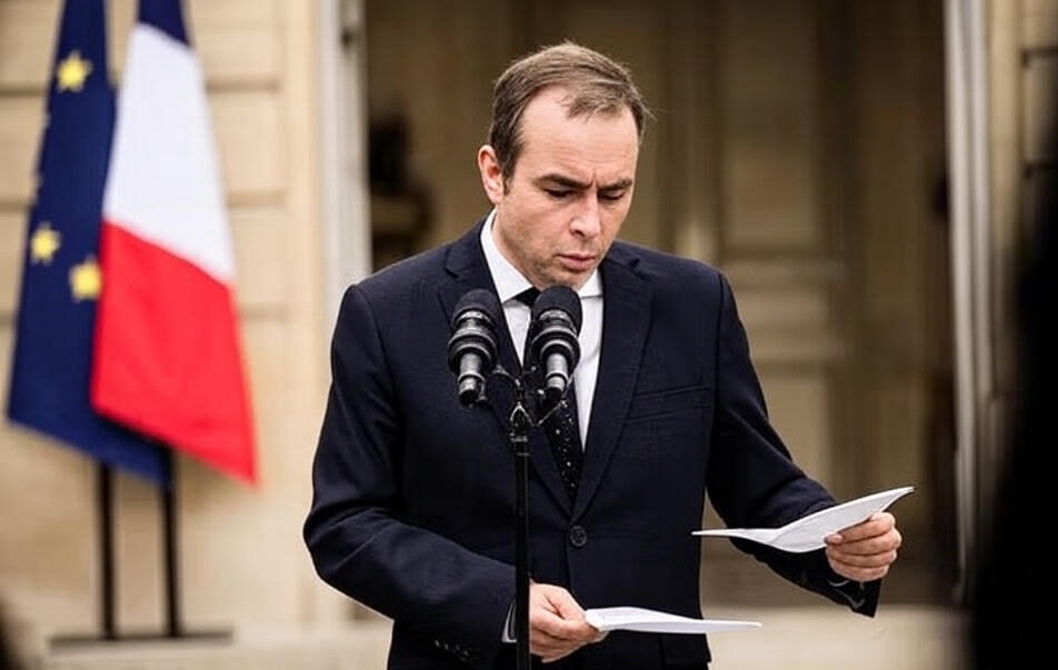 The image shows an individual dressed in a dark suit and tie, standing at a podium with two microphones, holding and reading from sheets of paper. The background features the French flag and a historic building entrance, suggesting an official or governmental setting.