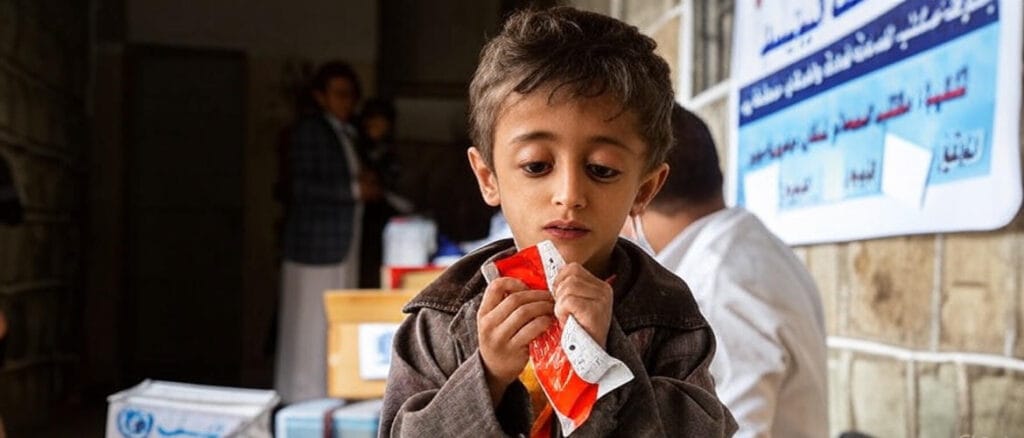 The image depicts a young child with dark hair, wearing a brown jacket, holding and eating a red and white packaged food item. The child is standing in a room with stone walls, surrounded by several people in the background. There are boxes with a logo and Arabic text, as well as a banner with Arabic writing, suggesting a humanitarian aid or relief distribution setting.