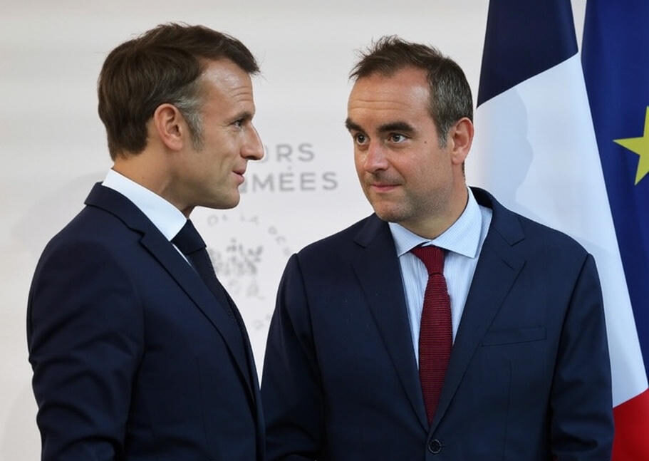 The image depicts two individuals dressed in formal dark suits, standing side by side in front of a backdrop featuring the text "MINISTÈRES" and the French flag. One individual is wearing a white shirt with a dark tie, while the other is wearing a light blue shirt with a red tie. The setting appears to be an official or governmental event.