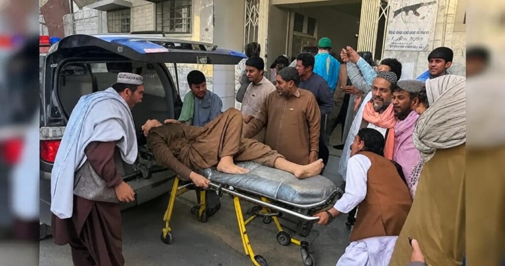 The image depicts an emergency medical scene where a group of individuals is transferring an injured or ill person on a yellow stretcher from an ambulance to a medical facility. The person on the stretcher is lying down, covered partially with a blanket, and appears to be receiving assistance. Several men, dressed in traditional attire and medical uniforms, are surrounding the stretcher, some assisting with the transfer and others observing. The ambulance, with its rear door open, is parked in front of a building with a sign featuring an image of a rifle and text in a local language. The scene takes place on a paved area outside the facility, with the group actively engaged in the process.