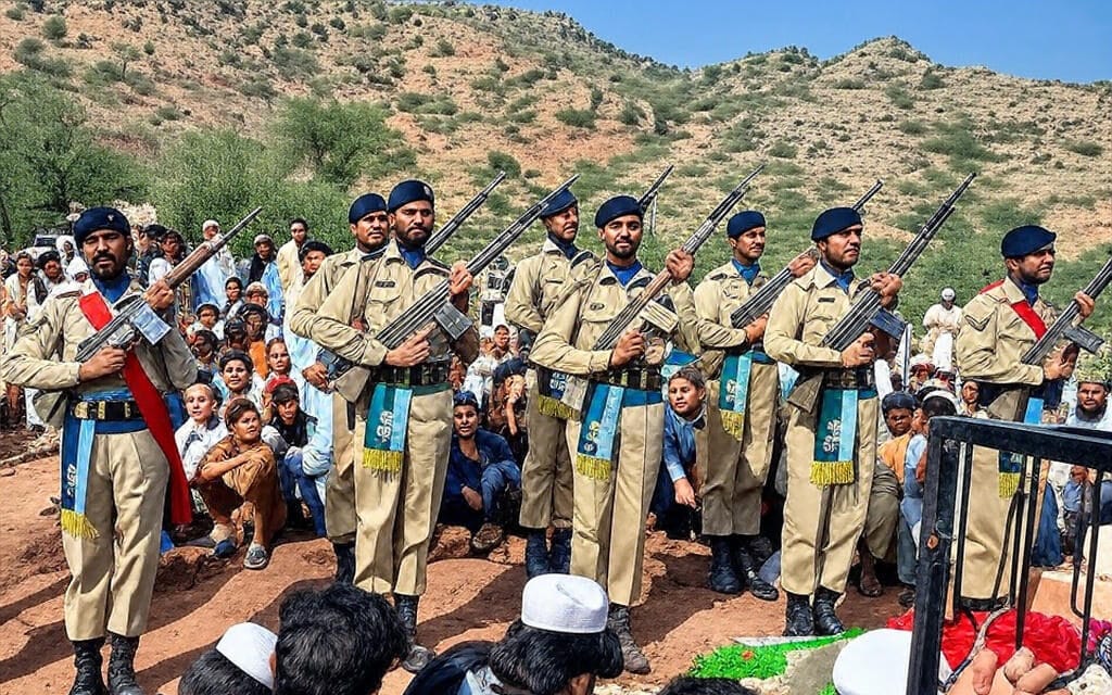 The image depicts a group of uniformed personnel standing in a line, holding rifles with bayonets fixed, in a formal military formation. They are dressed in khaki uniforms with blue berets, blue sashes, and decorative elements, suggesting a ceremonial context. In the foreground, a crowd of civilians, including men and children, is seated on the ground, observing the formation. The setting is an outdoor area with a mountainous landscape and sparse vegetation in the background, indicating a rural location. A floral arrangement and a metal structure are visible in the lower right corner, possibly part of a memorial or tribute site.