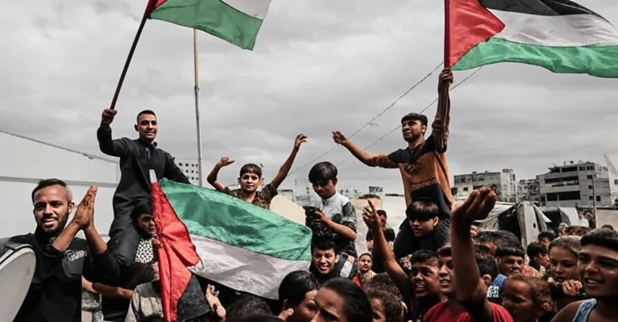 The image shows a large group of people, including adults and children, gathered closely together and raising their hands in a celebratory or defiant gesture. Several individuals are holding and waving Palestinian flags, which feature red, green, white, and black colors. The scene appears to take place outdoors, with a backdrop of buildings and a cloudy sky, suggesting an urban environment. The atmosphere conveys a sense of unity and strong emotion, likely in the context of a protest or demonstration.