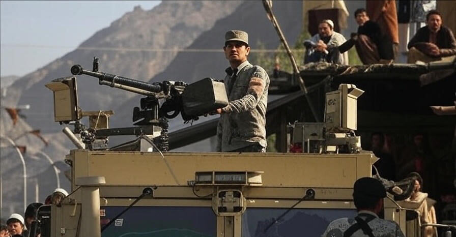 The image depicts a military scene featuring a soldier standing on top of an armored vehicle equipped with a mounted machine gun. The soldier is dressed in a camouflage uniform and cap, actively operating the weapon. The vehicle is positioned in a mountainous area with rugged terrain in the background. Several individuals, some in traditional attire, are observing from a raised platform or rooftop nearby. Additional personnel, including another soldier in the foreground, are present around the vehicle, suggesting a patrol or security operation. The setting appears to be a rural or semi-urban location with a mix of natural and man-made elements.