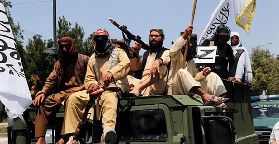 Armed individuals with Taliban flags riding on a military vehicle, holding weapons and displaying banners, set against a backdrop of trees and a clear sky.