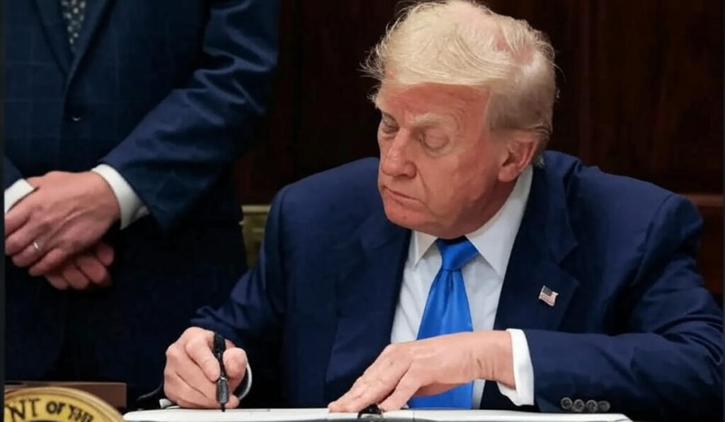 The image depicts an individual in a dark blue suit with a blue tie, seated at a desk and signing a document with a pen. The individual has light-colored hair and is wearing a small American flag pin on the lapel. Another person in a similar dark suit stands to the left, with hands visible. The desk features a seal with the text "DEPARTMENT OF THE" partially visible, indicating a formal or governmental setting. The image is credited to AFP.