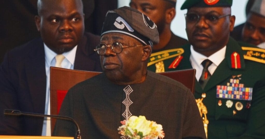 Nigerian President Bola Ahmed Tinubu, wearing traditional attire and glasses, sits sternly at a formal event flanked by a suited aide and a decorated military officer, with a bouquet of flowers on the table in front of him.