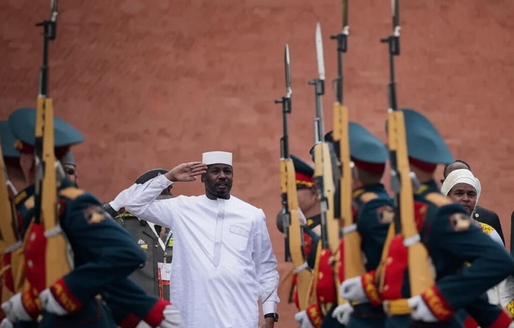 A man in traditional white attire and cap salutes while walking past a formation of soldiers in ceremonial uniforms holding rifles with bayonets fixed, against a red brick wall backdrop, during a formal military parade or inspection in Mali.