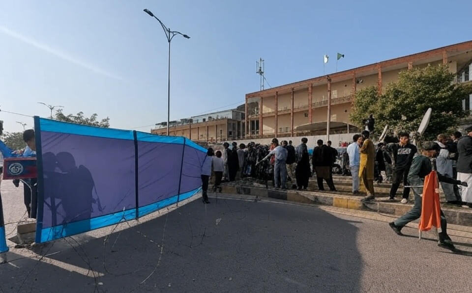 Security personnel erect blue tarpaulin screens and barbed wire to shield the site of the suicide bombing outside the district court complex in Islamabad, as journalists, lawyers, and onlookers gather under a clear sky with Pakistani flags visible on the building, capturing the tense aftermath of the November 11, 2025 attack.
