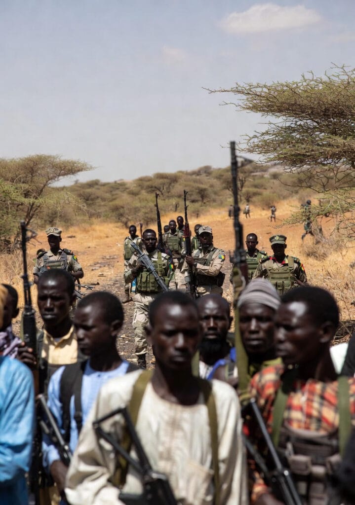 A group of armed soldiers in camouflage uniforms, some wearing tactical vests and carrying rifles, patrol a dusty rural path in the arid Sahel landscape of Mali; in the foreground, local civilians in traditional attire walk alongside them, while a military vehicle follows and displaced families gather near makeshift tents in the background, illustrating the intersection of security operations and humanitarian displacement amid the ongoing crisis.