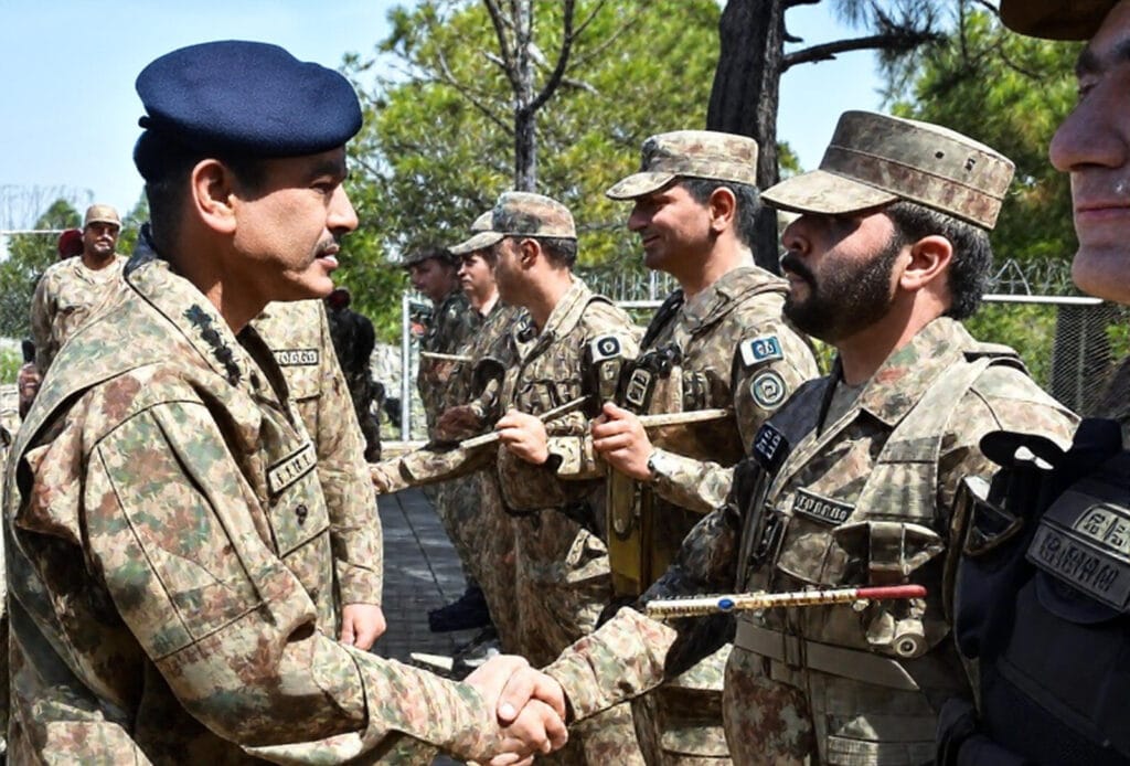 Pakistan Army Chief General Asim Munir, wearing a dark beret and camouflage uniform, shakes hands with a bearded soldier in full combat gear during an outdoor military ceremony, with a line of troops standing at attention behind them against a backdrop of trees and barbed wire fencing.