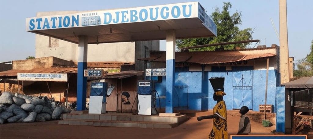 A deserted gas station named "Station Djebougou" in Mali stands empty under a blue and white canopy, with two fuel pumps covered in protective wraps; sacks of goods are piled nearby, while a woman in traditional yellow patterned dress carrying a load on her head walks past with a child, against a backdrop of modest buildings and trees, symbolizing the fuel shortages caused by the JNIM blockade.