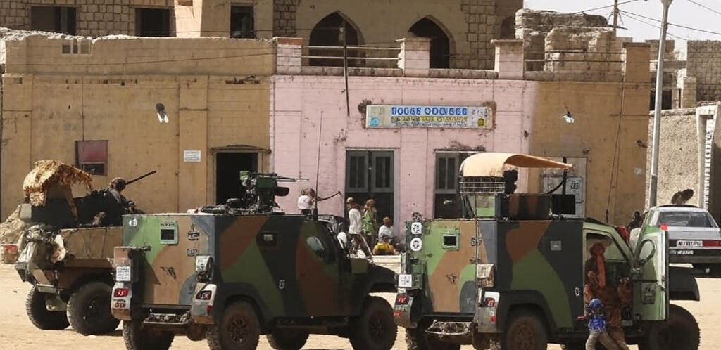 Armored military vehicles with mounted machine guns are parked in a dusty street in Timbuktu, Mali, in front of traditional mud-brick buildings; soldiers stand nearby while local civilians, including children, observe or walk past a pink storefront labeled "Bocar Couture," illustrating the tense security environment amid JNIM threats and the ongoing fuel blockade crisis.
